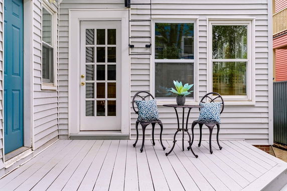 Rear view of a house with glass panel door and two windows, featuring a small deck and patio furniture.