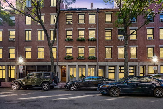 Front view of a multi-story brick building with illuminated windows and parked cars in front.