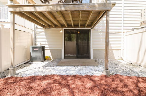 Rear view of a house with a wooden deck above, sliding glass doors, and an air conditioning unit.