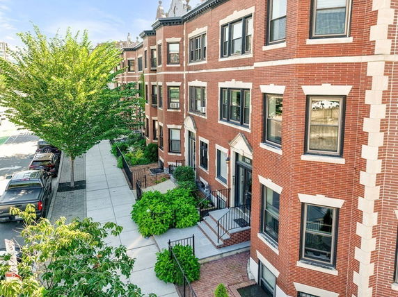 Front view of a multi-story brick residential building with adjacent sidewalk and greenery.
