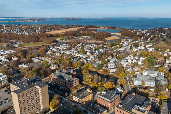 Aerial panoramic view of a coastal town with residential and commercial buildings, surrounded by water bodies and greenery.