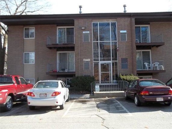 Front view of a three-story brick apartment building with balconies and a parking area.