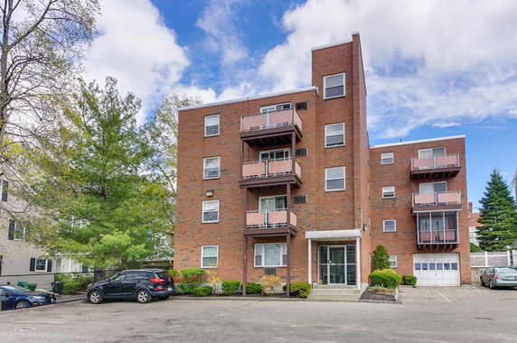 Front view of a multi-story brick apartment building with balconies and a parking area.