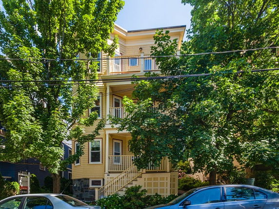 Front view of a multi-story yellow house with balconies, partially obscured by trees.