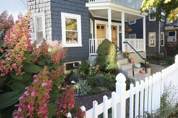 Front view of a house with a small porch and wooden steps, surrounded by a white picket fence and garden.