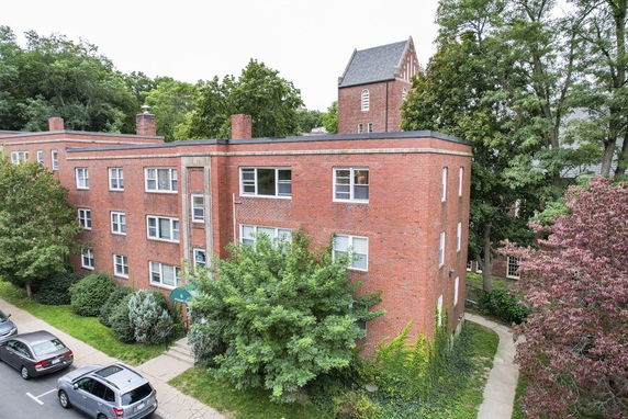 Front view of a multi-story brick apartment building with windows and surrounding trees.