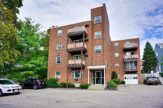 Front view of a multi-story brick apartment building with balconies and parking area in front.
