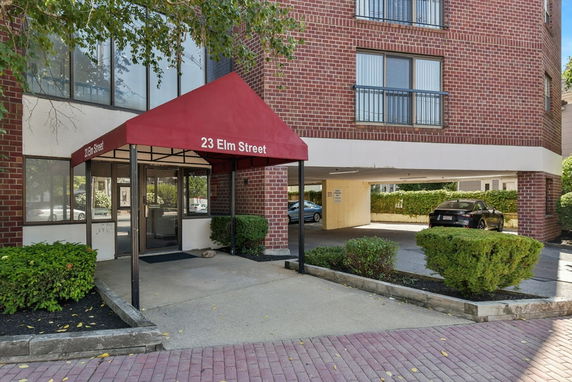 Front view of a brick building with a red awning entrance and adjacent parking area.