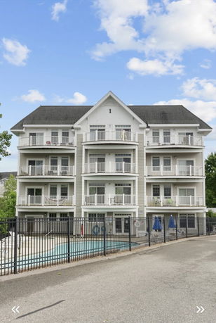 Front view of a multi-story building with balconies and a swimming pool in front.