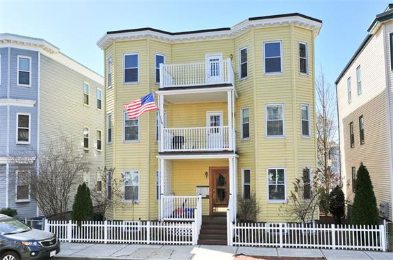Front view of a yellow three-story house with balconies and a white picket fence.