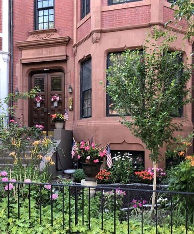 Front view of a red brick house with decorative windows and a wooden door, surrounded by a small garden.