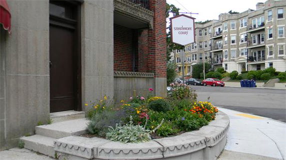 Entrance of a brick apartment building with signage and flower bed.