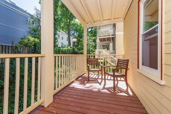 Wooden balcony with chairs, adjacent to a house, overlooking surrounding buildings and greenery.