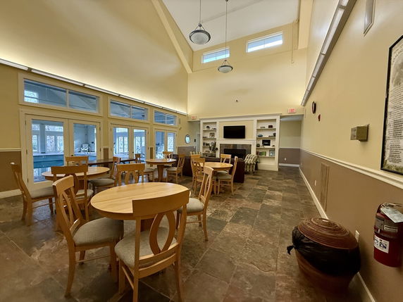 Interior view of a clubhouse with round tables and chairs, a fireplace, and large windows.
