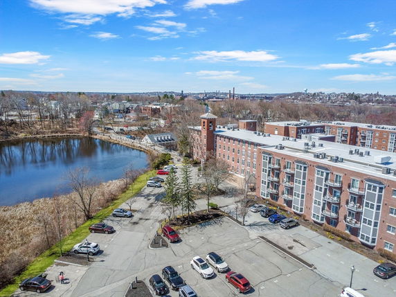Aerial view of a multi-story brick building with balconies, adjacent to a parking area and a pond.