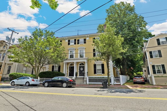 Front view of a multi-story yellow house with white trim and a fenced front yard.