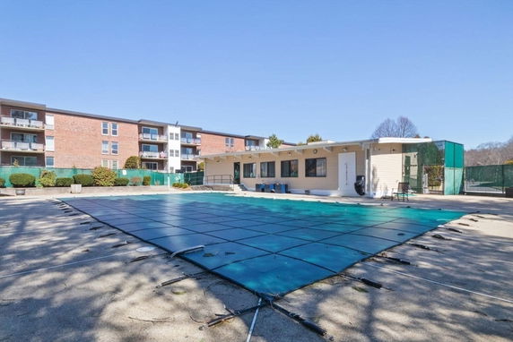 Outdoor swimming pool with a cover next to a small building and surrounded by apartment buildings.