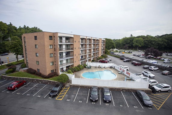View of a mid-rise apartment building with a parking lot and a swimming pool.