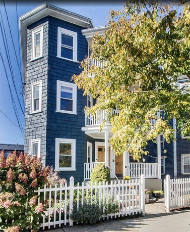 Front view of a multi-story house with blue siding and white trim, featuring a white picket fence and balconies.