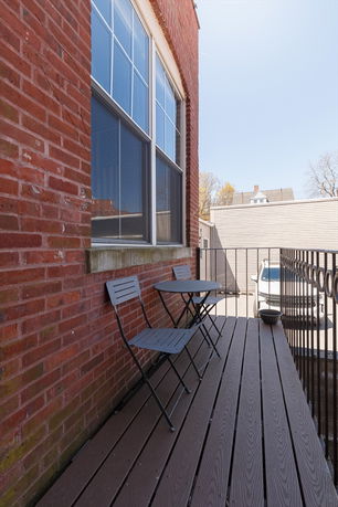 Outdoor balcony with chairs and a table, adjoining a brick building, overlooking a small parking area.