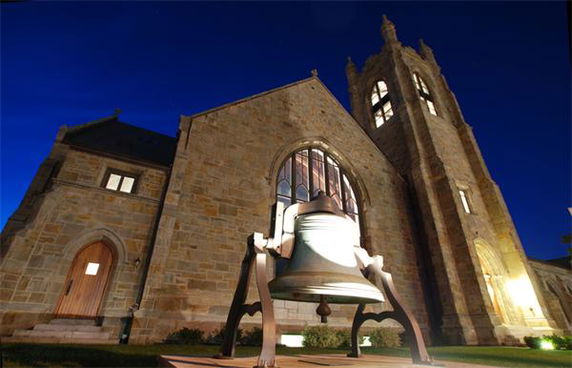 Front view of a stone building with a large bell displayed in the foreground.