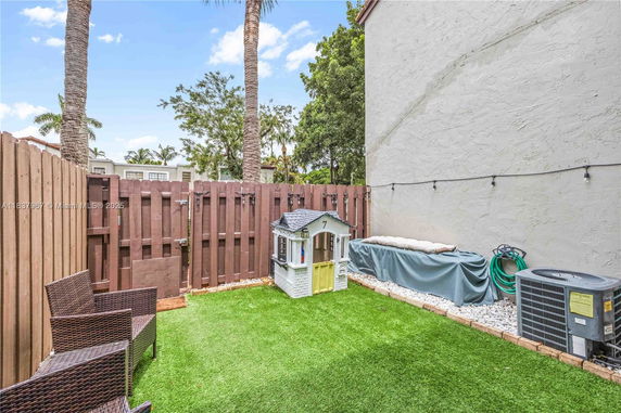 Fenced backyard with artificial grass, a small playhouse, chairs, and a side wall of a building.