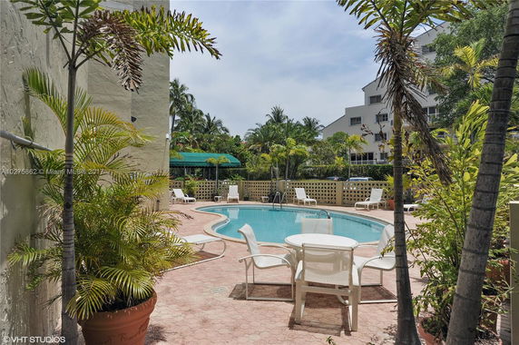 Outdoor swimming pool with lounge chairs and a patio table, surrounded by tropical plants and nearby buildings.