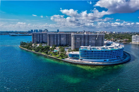 Aerial view of a waterfront building complex with curved architecture surrounded by water, with other residential buildings in the background.