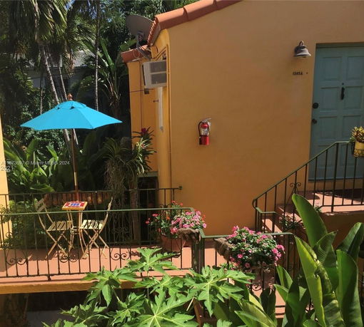 Exterior view of a small house with a yellow wall, blue door, and outdoor staircase leading to a patio with a blue umbrella.