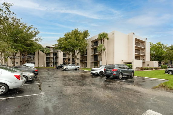 A multi-story apartment building with a parking lot in front, featuring multiple cars parked in marked spaces.