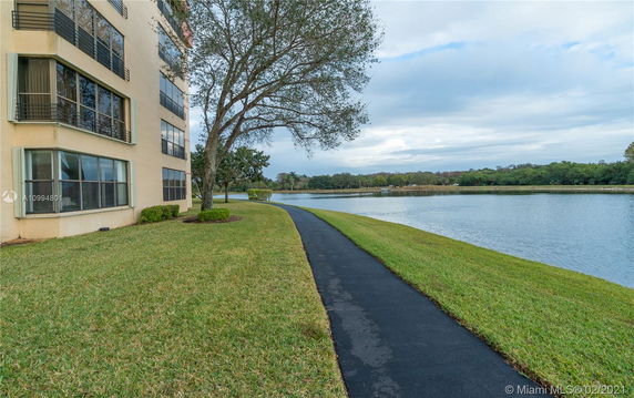 Side view of a multi-story building near a lake, with a walking path and grassy area.