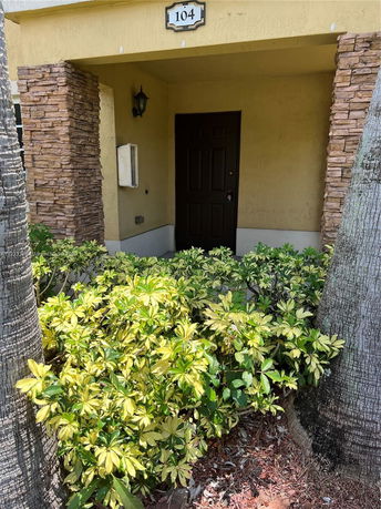 Front entrance of a building with a door labeled 104, stone pillars, and surrounding plants.