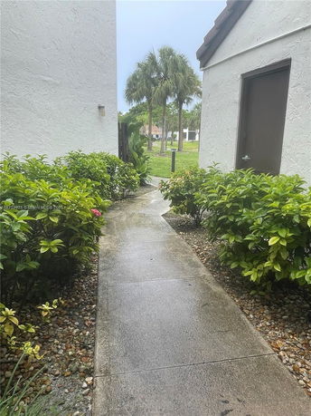 View between two buildings with a path, surrounded by shrubs, leading to an open grassy area with palm trees.