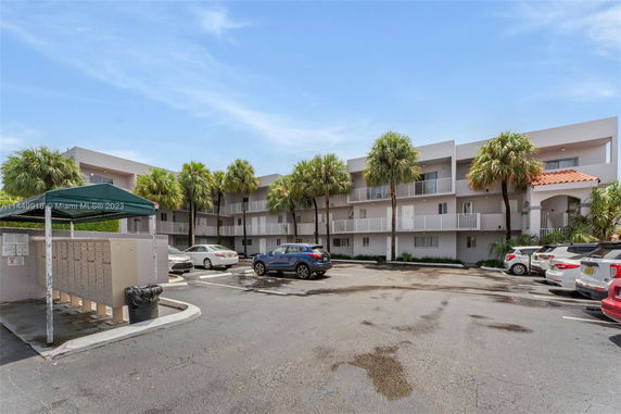Parking area in front of a multi-story apartment building with balconies and palm trees.