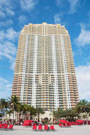Front view of a tall residential building with multiple balconies and a beach area with red lounge chairs.