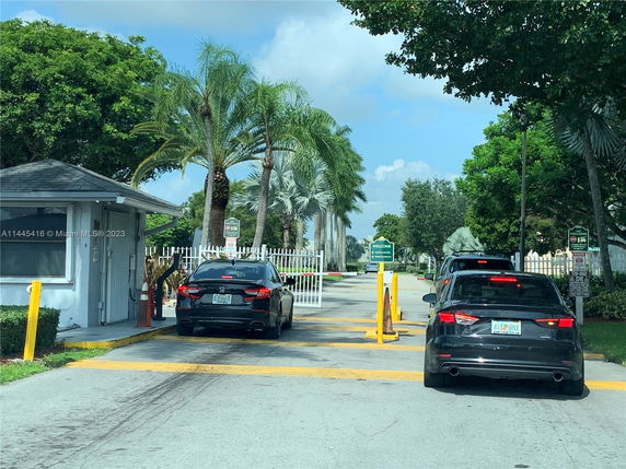 Entrance to a parking area with a security booth and cars queued at the gate.