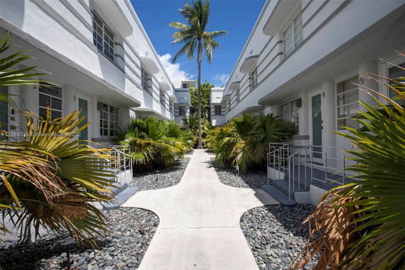 Front view of a multi-unit residential building with a central walkway and surrounding tropical plants.
