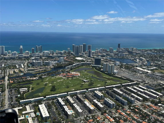 Aerial cityscape with numerous buildings, a large golf course in the foreground, and the ocean in the background.