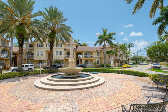Front view of three-story residential buildings with beige and white exterior, featuring balconies and surrounded by palm trees and a central fountain.