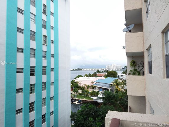 View between two buildings, one with a light beige facade and the other painted in blue and white stripes. Background includes a river and distant high-rise buildings.