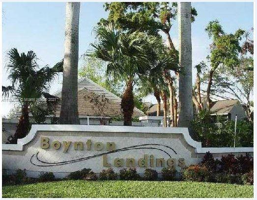 Front view of a residential area entrance sign with palm trees and houses in the background.