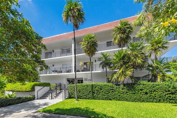 Front view of a three-story building with balconies and palm trees in the foreground.