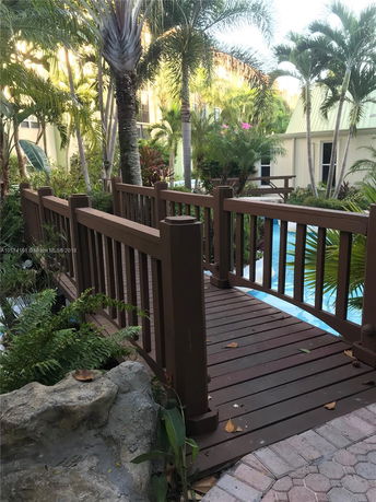 Wooden bridge surrounded by lush greenery and palm trees with buildings in the background.