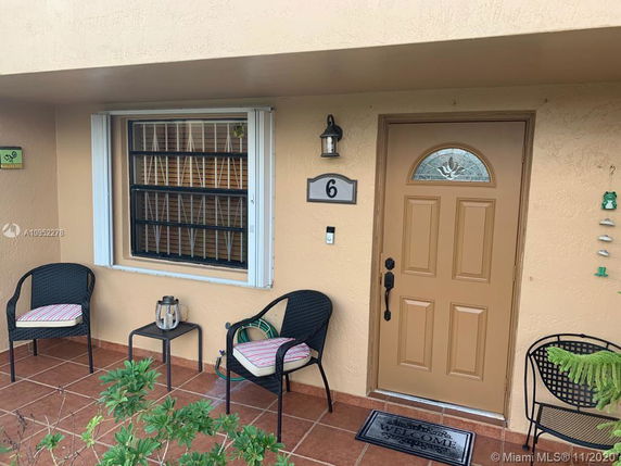 Front view of a house entrance with a brown door, window with bars, and patio chairs.