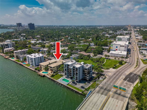 Aerial view of waterfront buildings with swimming pools and a main road nearby, situated along a body of water.