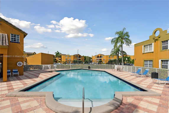 View of a rectangular swimming pool surrounded by residential buildings with palm trees nearby.