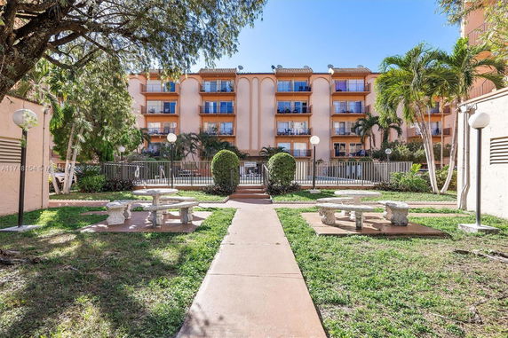 View of a multi-story apartment building with balconies and a landscaped area with stone tables and benches in the foreground.