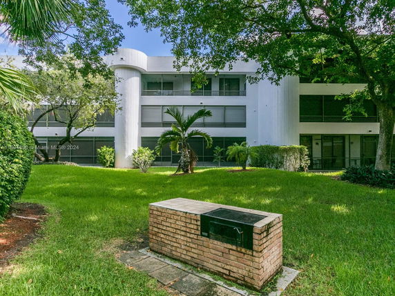 Front view of a modern multi-story building with large windows and a circular column. The building is surrounded by grass and trees.