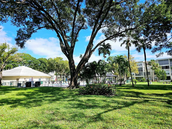 View of a residential area with a green lawn and trees, adjacent buildings, and a pool area visible.
