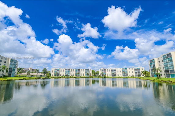 A row of multi-story residential buildings with a pond in the foreground under a partly cloudy blue sky.